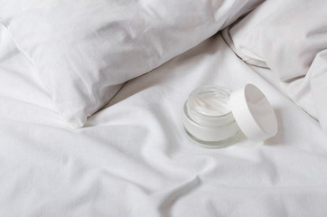 Fototapeta na wymiar Glass jar of face moisturizer cream, pillow, white sheet on the bed.Concept of morning beauty routine