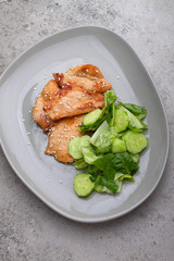 Fried chicken fillet in soy sauce on a plate decorated with sesame seeds and green salad, top view.