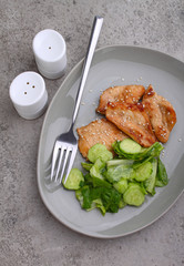Fried chicken fillet in soy sauce on a plate decorated with sesame seeds and green salad, top view.