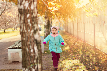 little girl 3 years in blue jacket, red pants, fur hat laughs and runs on the yellow foliage in autumn park