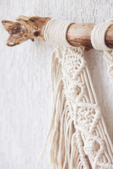 Fototapeta na wymiar Handmade macrame 100% cotton wall decoration with wooden stick hanging on a white wall. Macrame braiding and cotton threads. Details close up. Female hobby.