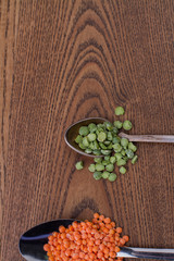 Teaspoon with bunch of green split peas. Tablespoon with lentils groats. Brown wooden background.