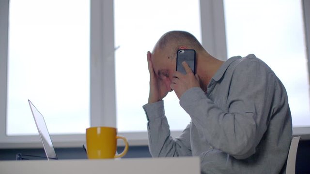 Side view of stressed man talking on smartphone at workplace. Exhausted businessman sitting at table with laptop and talking with interlocutor through phone. Economic crisis concept