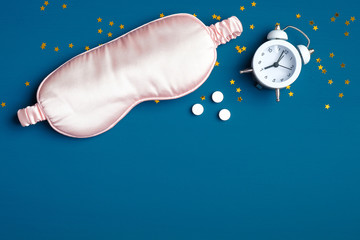 Pink sleep eye mask, alarm clock, sleeping aid on dark blue background. Flat lay, top view. Good night, sleep hygiene, insomnia treatment concept.