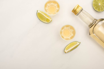 top view of golden tequila in bottle and shot glasses with lime on white marble surface