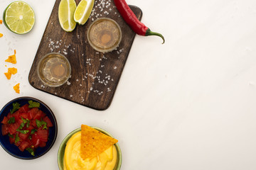 top view of golden tequila with lime, chili pepper, salt and nachos with cheese sauce near wooden cutting board on white marble surface