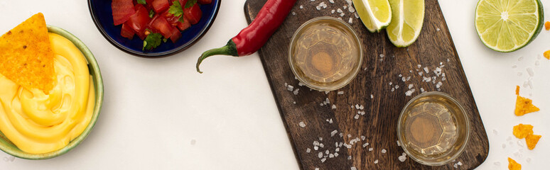 top view of golden tequila with lime, chili pepper, salt and nachos with cheese sauce near wooden cutting board on white marble surface, panoramic shot