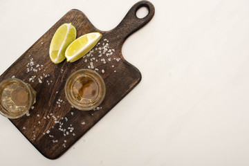 top view of golden tequila with lime, salt on wooden cutting board on white marble surface