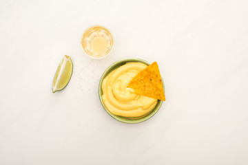 top view of golden tequila with lime and bowl with cheese sauce and nachos on white marble surface