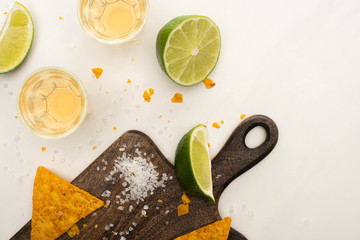 top view of golden tequila in shot glasses with lime, salt and nachos on wooden cutting board on white marble surface