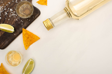 top view of golden tequila with lime, salt and nachos near wooden cutting board on white marble surface