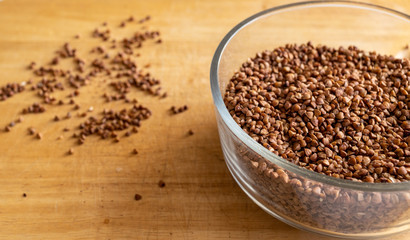 raw buckwheat in a transparent deep bowl. seeds are scattered carelessly nearby. kitchen table. daylight