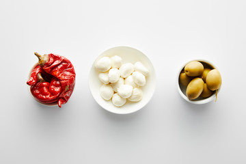 Top view of bowls with olives, marinated chili peppers and mozzarella on white background