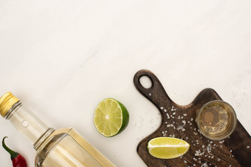top view of golden tequila with lime, chili pepper, salt on wooden cutting board on white marble...