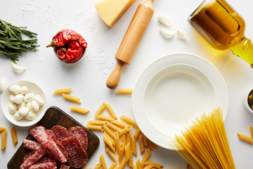 Top view of plate, bottle of olive oil, meat platter, rolling pin, pasta and ingredients on white