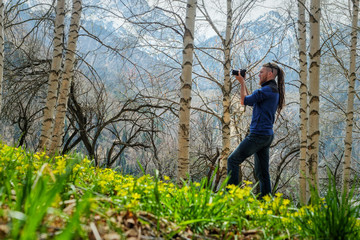 A man photographer stands on the trail in the spring birch forest. The guy holds a camera in his hand. View from the back. Fresh young grass and wildflowers. Alma-Ata's region. Kazakhstan.