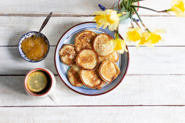 Fritters in blue plate, honey with spoon, cup of coffee with lemon, daffodile flowers bouquet on white wooden background