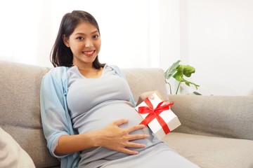 Beautiful Asian mother sitting on sofa at home looking and touching her baby bump with a gift box wrapped with red ribbon in her hand. Happy mom smiling and expecting baby with cute present concept