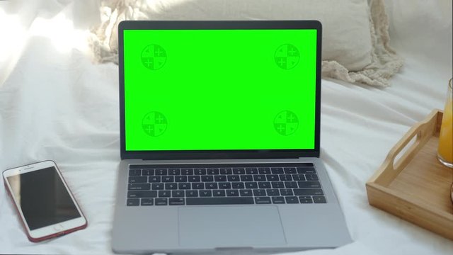 Mock-up Laptop Computer with Chroma Key Green Screen Set on Blanket in Home Office. A Glass of Orange Juice and a Croissant Placed near the Computer on a Tray. Concept of Working in Bed from Bedroom. 
