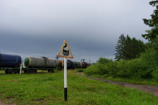 Road sign "Railway crossing without barrier" on the background of a train with oil tanks and a country road