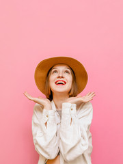 Surprised girl in a checkered face stands on a pink background, looks up at copy space 6 spreads her arms to the sides, wearing a sun hat and summer light clothing. Vertical.