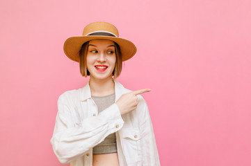 Happy lady in a light wear and hat stands on a pink background, looks away with a smile on face and points her hands away at copyspace. Smiling girl points fingers to the empty space