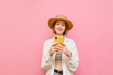 Happy girl in light clothing and sun hat stands on a pink background and uses a smartphone with a smile on her face. Lady uses the internet on her phone and smiles. Isolated.