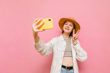 Closeup portrait of attractive girl in summer clothes and hat takes selfie on pastel pink background, looks into camera and smiles. Happy tourist girl makes photo on smartphone. Isolated.