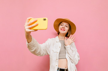 Happy girl in a hat and light summer clothes makes a video call on a pink background, looks at camera of a smartphone and greets her palm, shows a gesture of "hello. Lady in a hat calls on the video