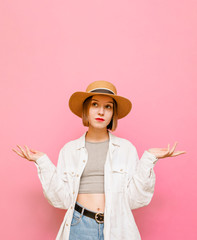 Funny woman in a hat looking puzzled up with raised arms from the bewildered hands on a pastel pink background. Portrait of puzzled girl in light clothing spreads her hands, isolated.