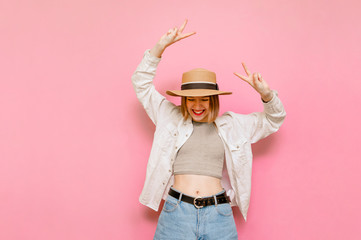 Happy woman in hat and summer clothes rejoices with raised hands on pink background, looks down at copy space and shows a gesture of peace.Joyful lady tourist isolated on pink background.