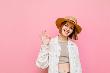 Pretty girl in hat and light summer clothes showing gesture OK and looking at camera, isolated on...