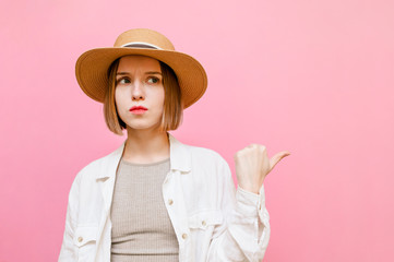 Serious girl in a hat and light clothing stands on a pink background,looks away and points a finger at copy space.Lady tourist in summer clothes with.Pensive sad face shows finger aside on empty space