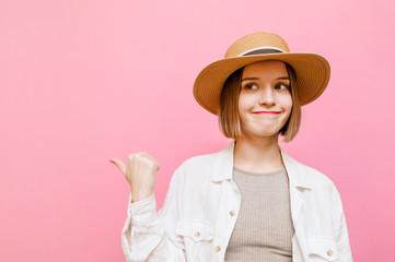 Funny girl in hat shows thumbs up on copy space and makes funny face,looks away at blank space,wears vacation clothes.Cheerful lady with a sad funny face points her finger to the side.Pink background