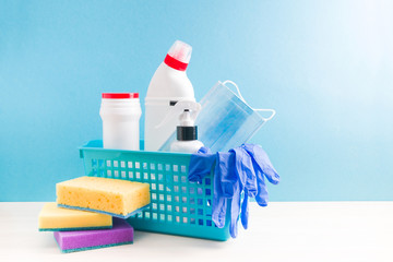 in a blue plastic basket are different plastic bottles with cleaning products, rubber disposable gloves and a protective fabric mask on a blue background, cleaning sponges on the table