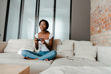 KYIV, UKRAINE  APRIL 10, 2020: african american woman with crossed legs playing video game with joystick on sofa in living room