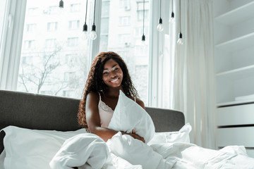 African american woman holding pillow, smiling and looking at camera on bed in bedroom