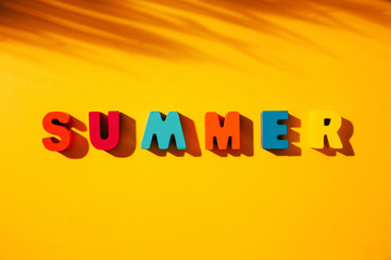 Flat lay composition with phrase SUMMER made of colorful wooden letters on top of yellow background, with palm leaf shadow next to it.