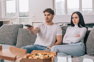 serious couple having pizza and watching tv with remote controller during self isolation at home