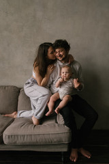 Young beautiful family mom and dad plays with toddler boy son in a living room. Family portrait in dark tones. Stylish minimalistic interior, family at home, tenderness, joy, fun, hugs.