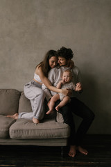 Young beautiful family mom and dad plays with toddler boy son in a living room. Family portrait in dark tones. Stylish minimalistic interior, family at home, tenderness, joy, fun, hugs.
