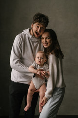 Young beautiful family mom and dad plays with toddler boy son in a living room. Family portrait in dark tones. Stylish minimalistic interior, family at home, tenderness, joy, fun, hugs.