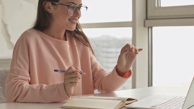 A smiling girl wearing glasses is writing something in her notebook using her laptop computer sitting at the table in the kitchen at home