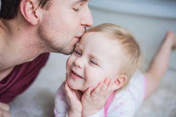 Blond-haired blue-eyed baby girl and young dad in the home interior. Dad kisses his little daughter.