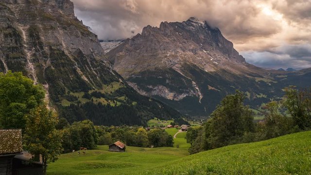 Time lapse of dark storm clouds moving over the Eiger mountain ranges in the Swiss Alps of Grindelwald Switzerland. The green farmland valley with alpine houses is dwarfed by the tall mountain peaks.
