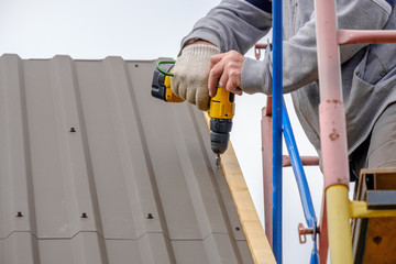 Men's hands in work gloves with a yellow screwdriver screw the roofing sheet to the roof of a...