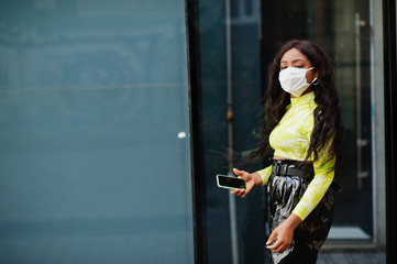 Naklejka premium African American woman posing with facial mask to protect from infections from bacteria, viruses and epidemics and hold mobile phone.
