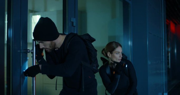 Couple of Caucasian male and female thieves breaking into house at night. Young man and woman in black costumes opening door and coming in somebody's home. Housebreakers concept.