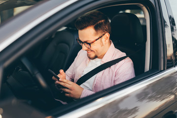 Side view through open window of a driver sitting in a car and looking in smartphone. A smiling happy young man with beard and stylish glasses