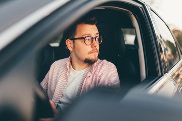 A young stylish man with good hairstyle and beard wearing glasses driving a car and looking out the open window